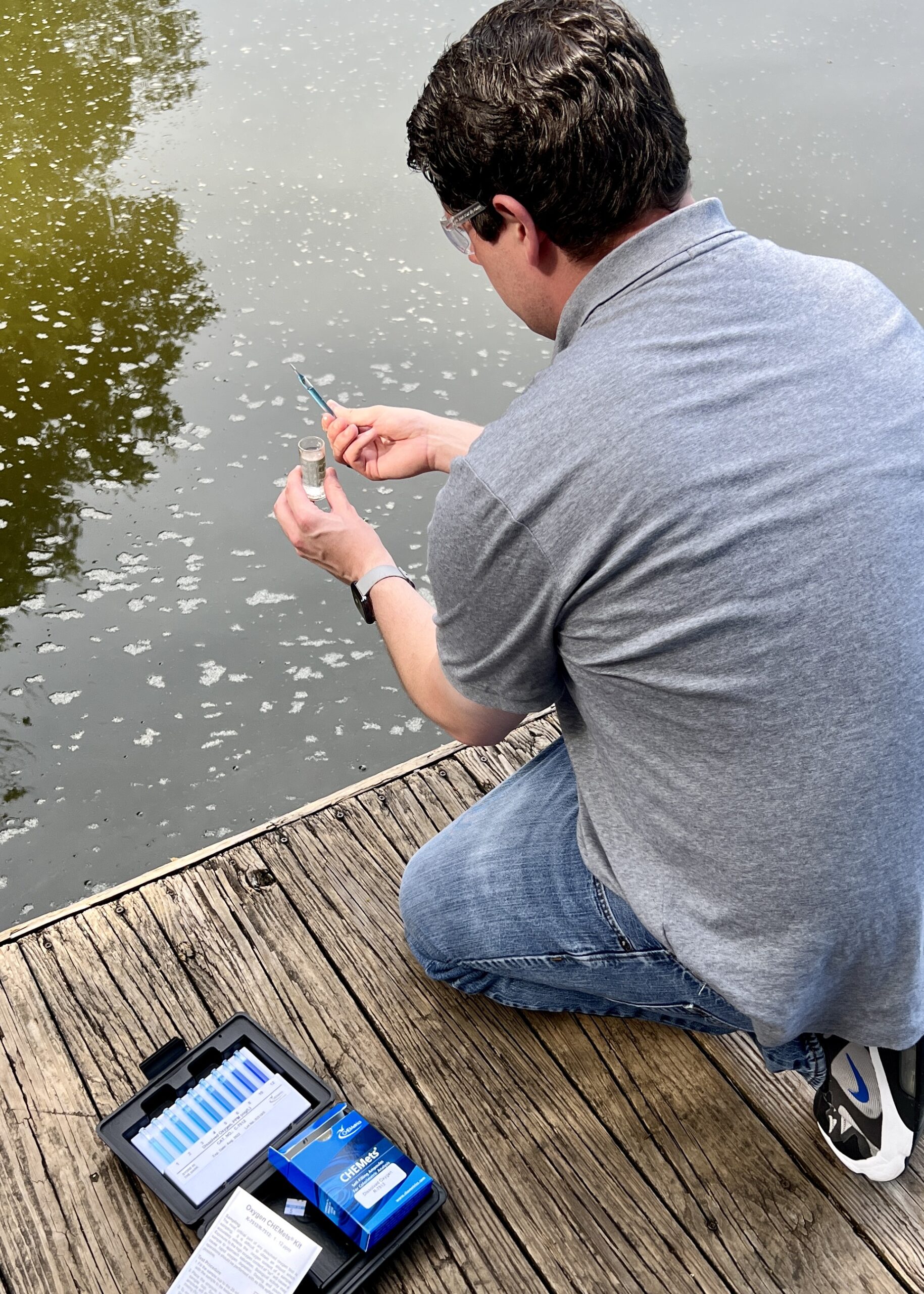 An operator tests lake water from a dock using a CHEMetrics dissolved oxygen test kit.